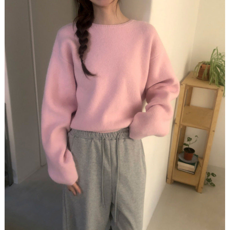 A person wearing a soft pink knit sweater with voluminous long sleeves and light gray sweatpants, standing indoors near a wooden shelf with small plants.
