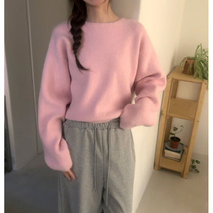 A person wearing a soft pink knit sweater with voluminous long sleeves and light gray sweatpants, standing indoors near a wooden shelf with small plants.