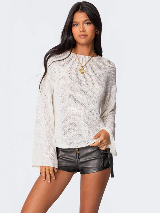 A woman modeling a cream-colored, textured knit long-sleeve top with flared bell sleeves, paired with black faux-leather shorts, against a solid light gray background.