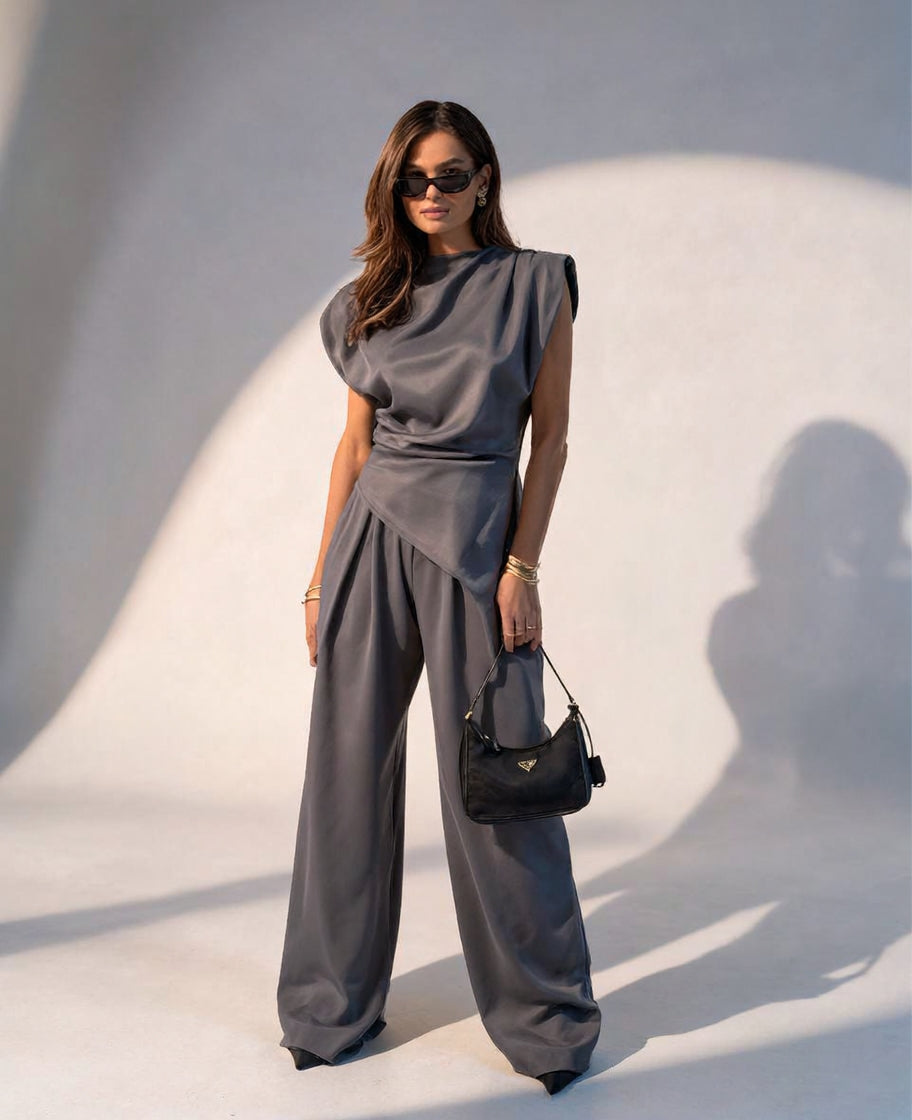 A woman models a gray draped sleeveless top with matching wide-leg pants, styled with a black handbag and sunglasses against a curved white wall with strong shadows.