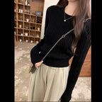A woman wearing a black cable-knit long-sleeve sweater and light beige pleated trousers, styled with a silver chain strap bag and a delicate silver necklace in an indoor setting.