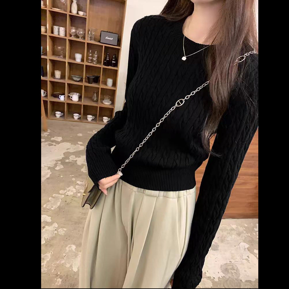A woman wearing a black cable-knit long-sleeve sweater and light beige pleated trousers, styled with a silver chain strap bag and a delicate silver necklace in an indoor setting.