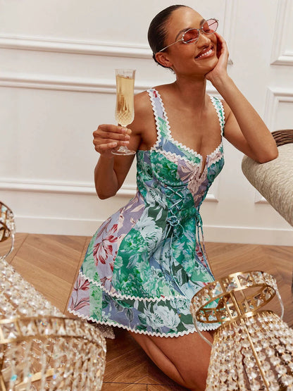 A woman wearing a colorful floral print mini dress and pink sunglasses holds a glass of champagne while kneeling on a wooden floor in an elegant room.
