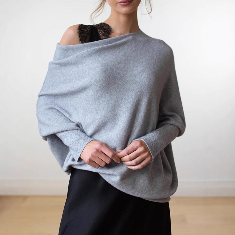 A woman wearing a light gray oversized knit sweater draped off the shoulders, paired with a black skirt, shown against a plain indoor wall.