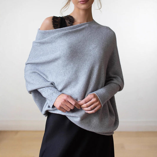 A woman wearing a light gray oversized knit sweater draped off the shoulders, paired with a black skirt, shown against a plain indoor wall.