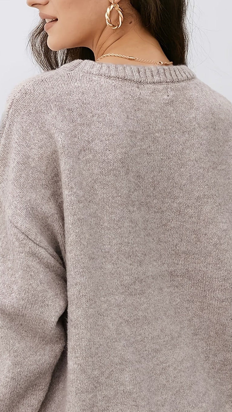 A woman wearing a light taupe knit sweater, shown from the back. The sweater has a soft, heathered texture and ribbed collar, cuffs, and hem, paired with white pants against a plain light gray background.