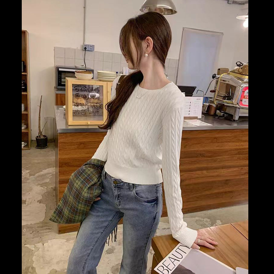 A woman wearing a white cable-knit long-sleeve sweater and blue jeans, standing in a cafe setting with a green plaid jacket draped over her shoulder.