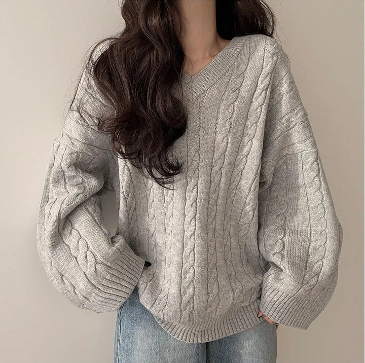 A woman wearing an oversized light gray cable-knit sweater with voluminous sleeves and light blue jeans, shown from the neck down against a plain beige wall.