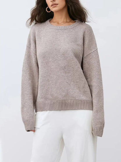 A woman wearing a light taupe knit crewneck sweater and white pants, shown from the nose down against a plain light gray studio background.