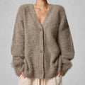 A person wearing a fuzzy, oversized V-neck cardigan in a warm taupe-brown color, paired with light beige pants, against a neutral gray background.