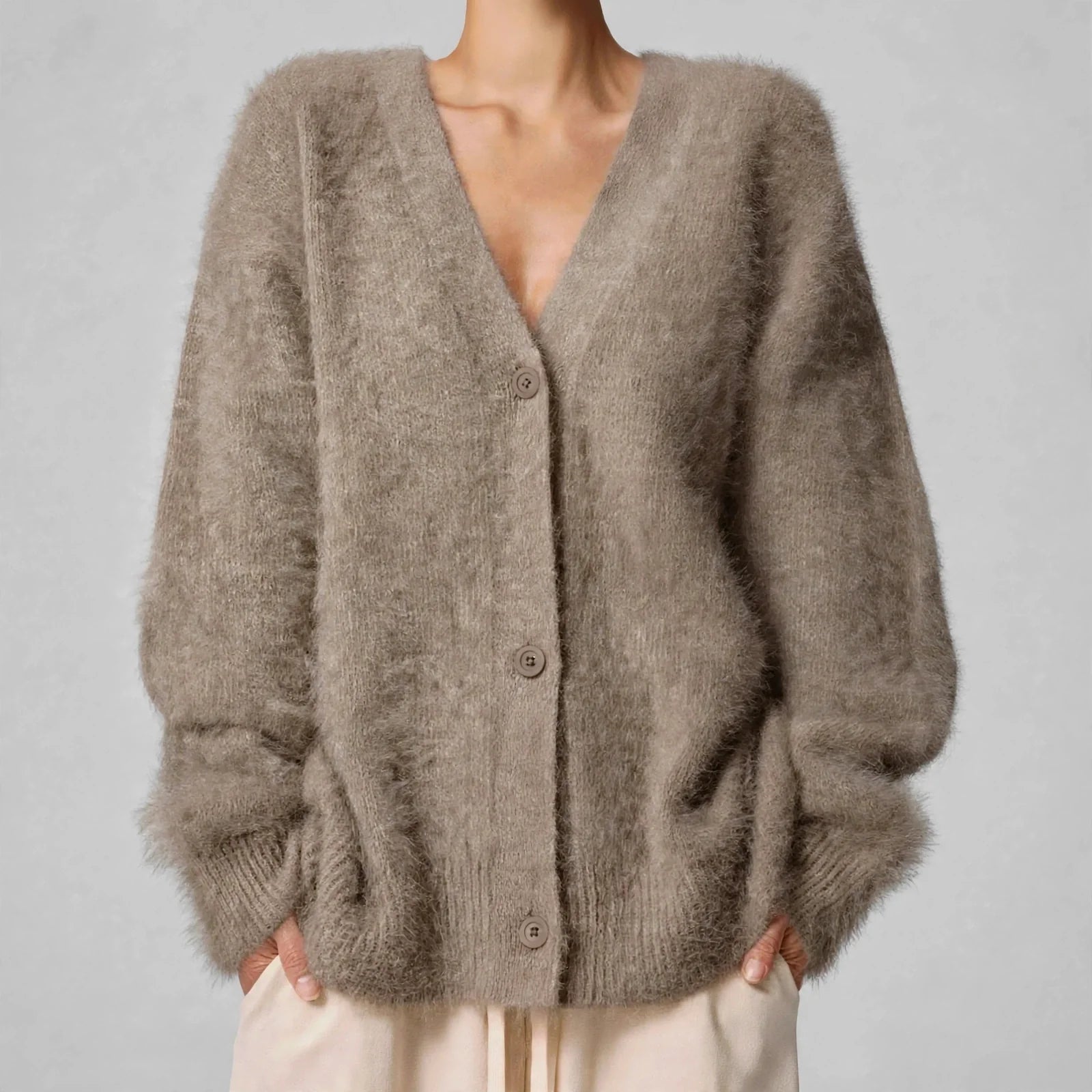 A person wearing a fuzzy, oversized V-neck cardigan in a warm taupe-brown color, paired with light beige pants, against a neutral gray background.