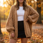 A woman wearing an oversized chunky knit cardigan in a warm camel brown, styled over a white t-shirt and a black mini skirt, standing in an autumn park with fallen leaves and soft golden light.