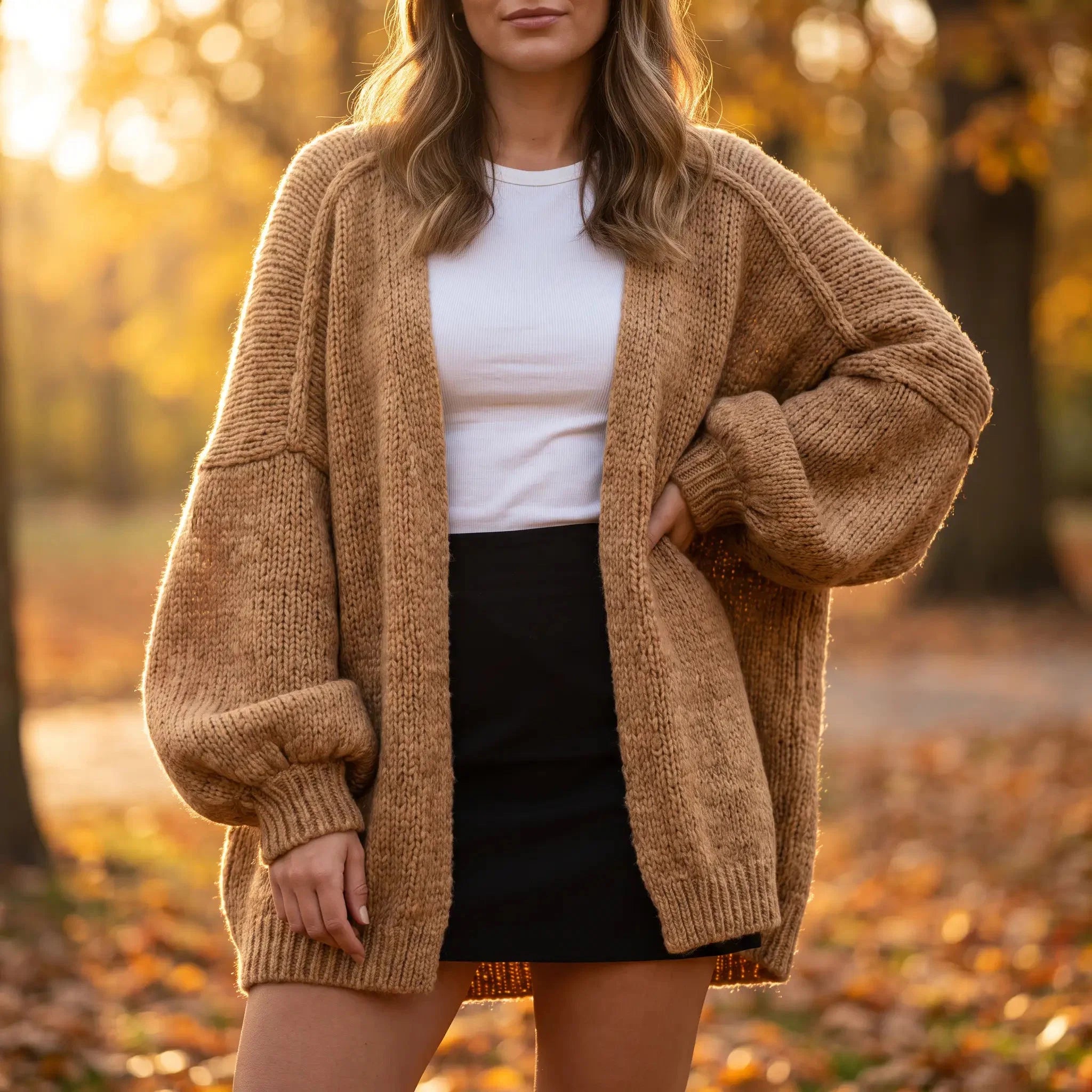 A woman wearing an oversized chunky knit cardigan in a warm camel brown, styled over a white t-shirt and a black mini skirt, standing in an autumn park with fallen leaves and soft golden light.